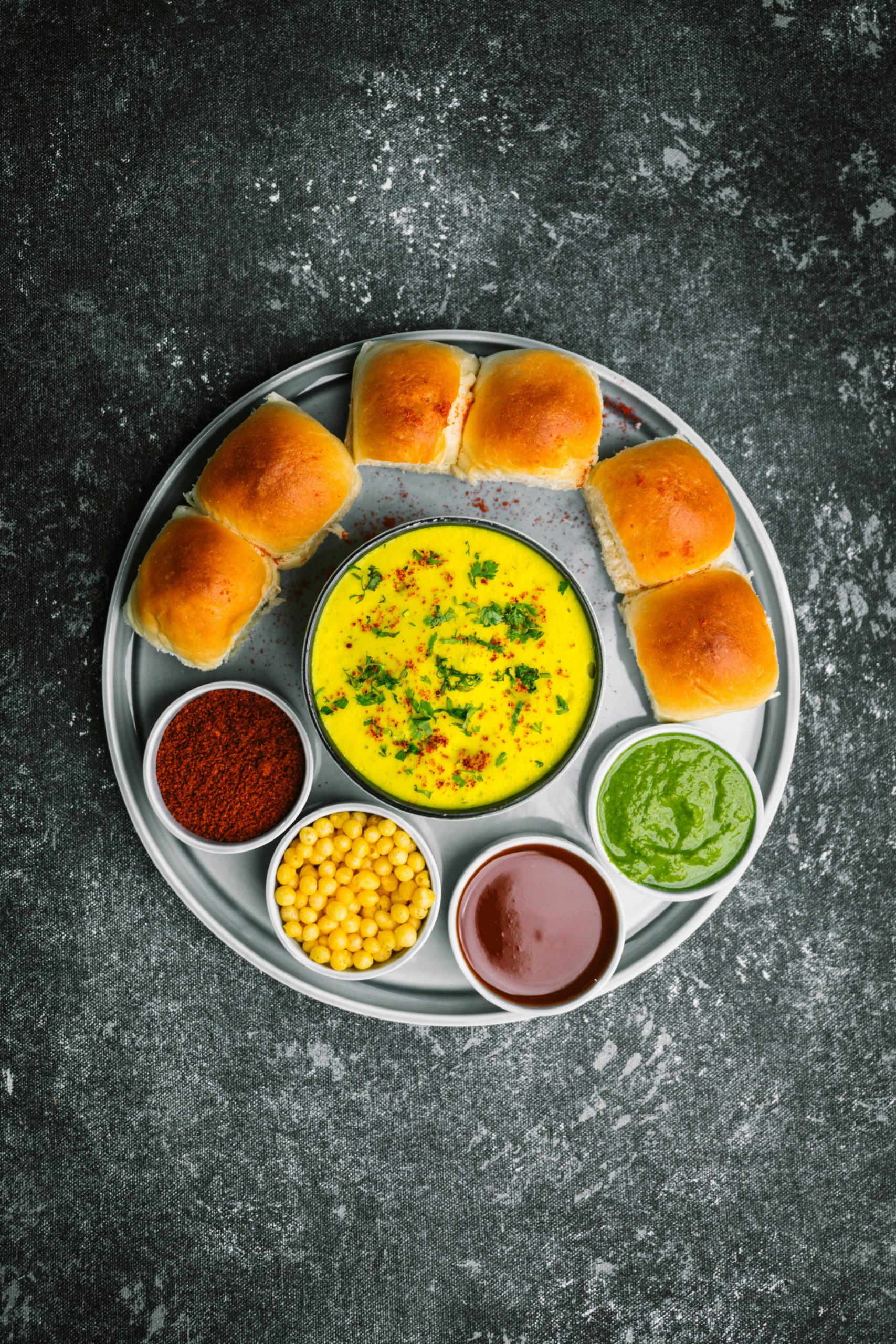 Top view of a colorful Indian pav bhaji meal on a stainless steel tray with assorted sauces.